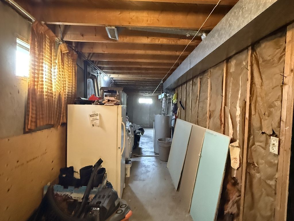 An unfinished basement corridor with open wooden ceiling joists, insulation showing behind bare wall studs, sheets of drywall leaning against the wall and a water heater at the far end