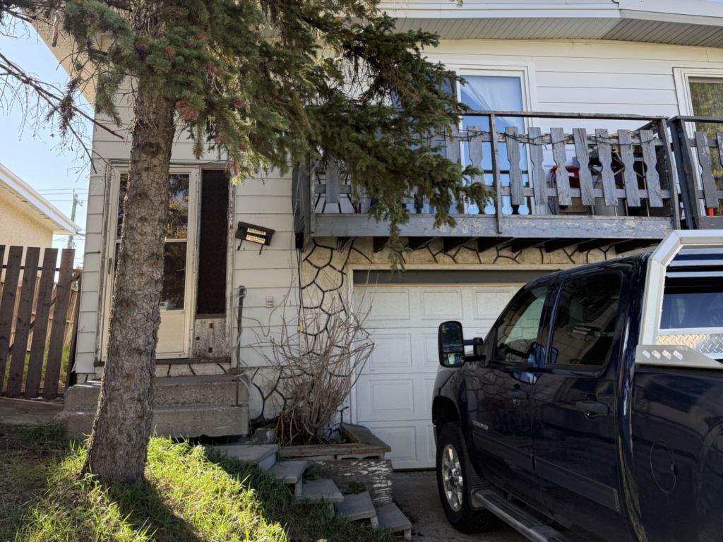 A white-sided house in Penbrooke Meadows SE Calgary with a weathered grey wooden balcony above a single garage, a tall spruce in front of the entry and a dark pickup truck in the driveway