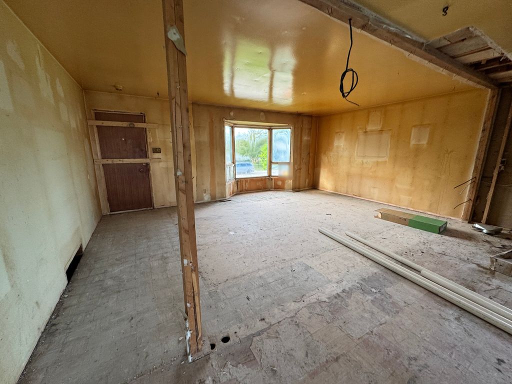 An empty room with yellow walls and a bay window, a brown door braced shut with two boards and a bare wooden post standing on a worn tile floor