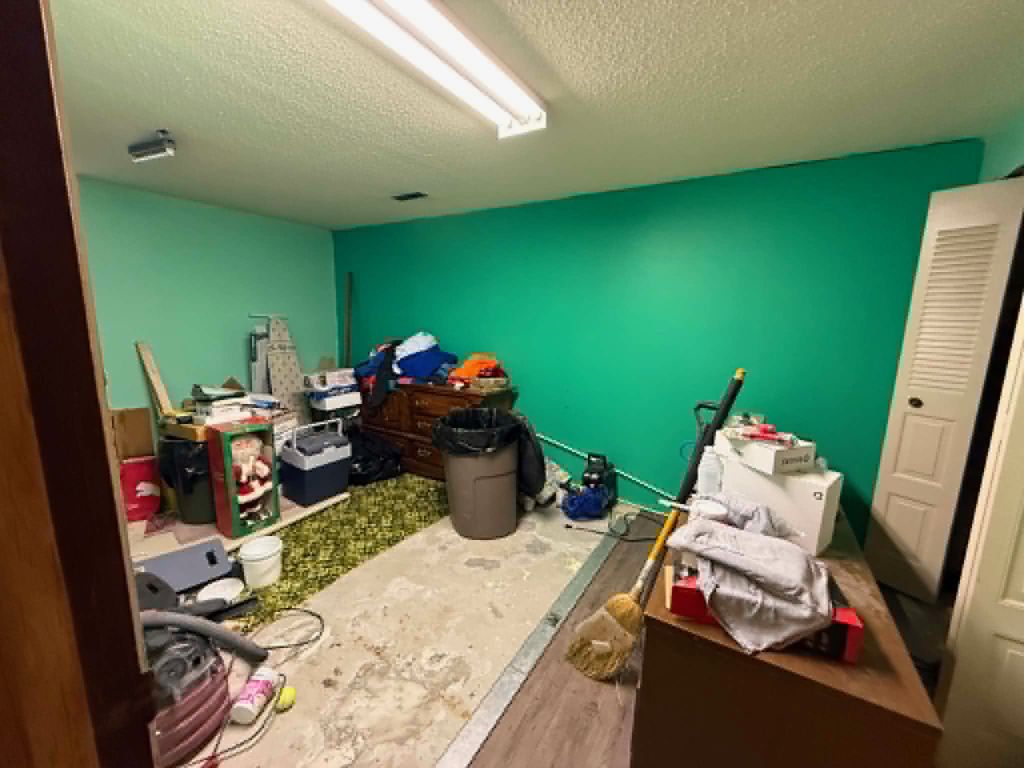 A room with teal walls and a fluorescent ceiling light, crowded with boxes, a dresser, a cooler, a garbage can and a broom, the floor covering partly torn up