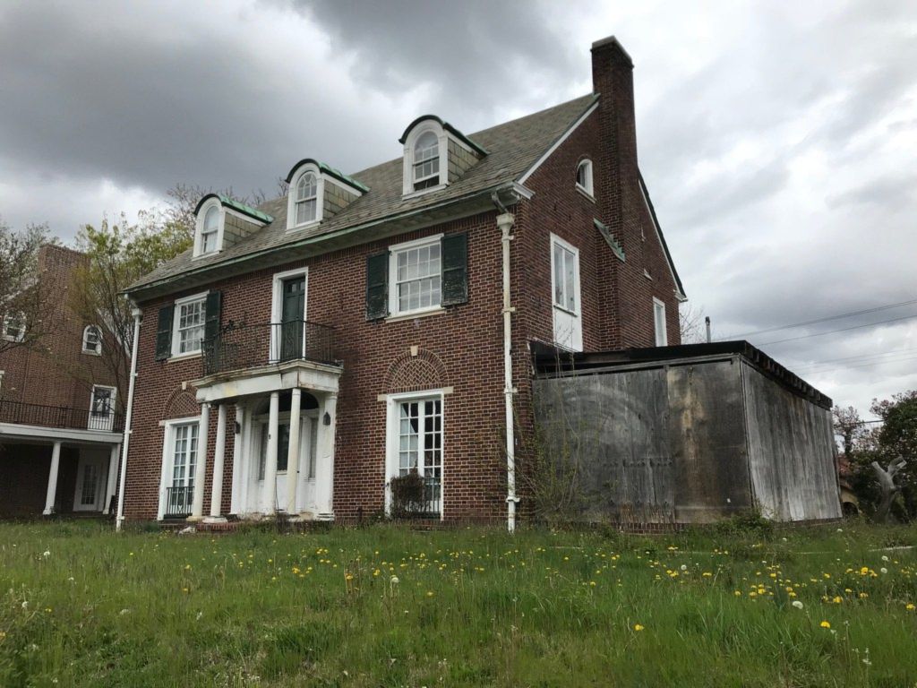 A large brick house with dormer windows and a columned entrance, dandelions coming up through the uncut lawn